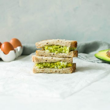 two egg and avocado sandwiches piled on top of each other with eggs and avocado in the background