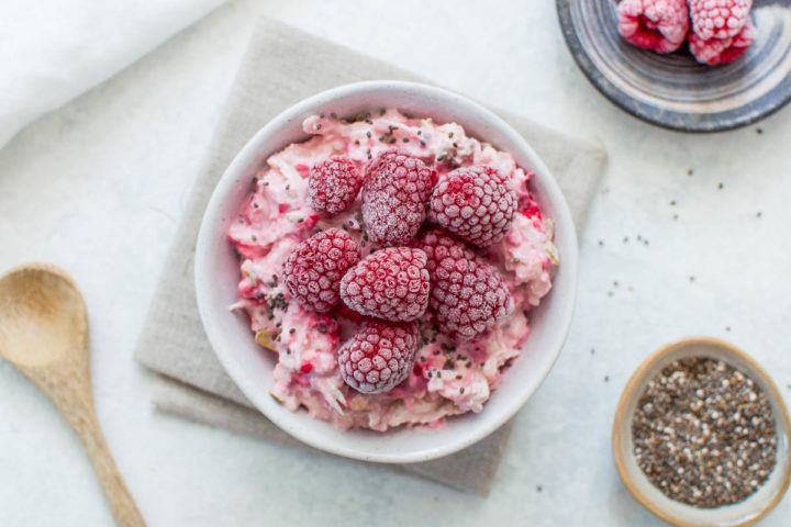 raspberry bircher in white bowl topped with frozen raspberries with wooden spoon to the left