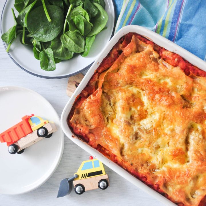 cheese and tomato lasagna in white baking dish with spinach salad and toy lorries on left hand side