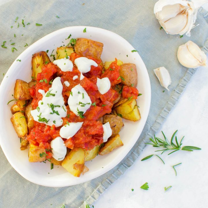 white bowl with patatas bravas sprinkled with fresh parsley with garlic cloves and fresh rosemary to the right