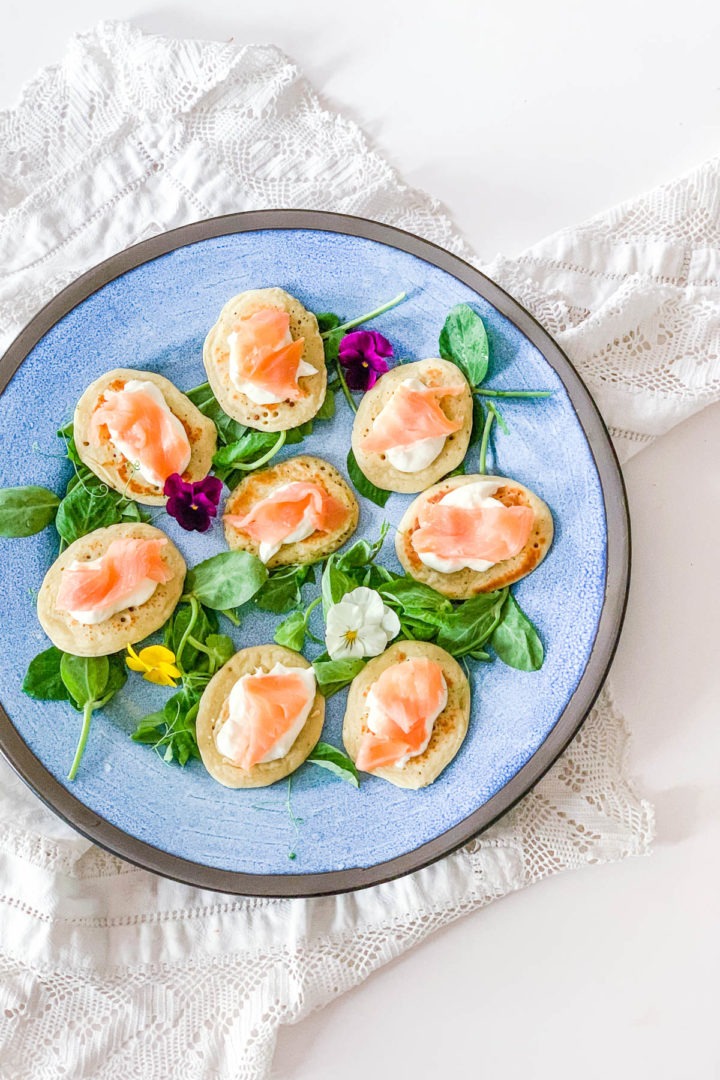 blinis topped with smoked salmon on blue plate decorated with edible flowers on white background