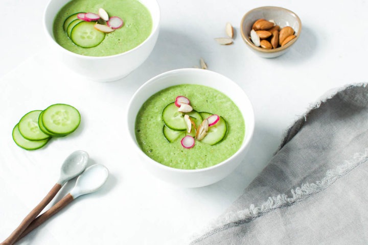 angled view of two white bowls of green cucumber gazpacho with bowl of almonds behind
