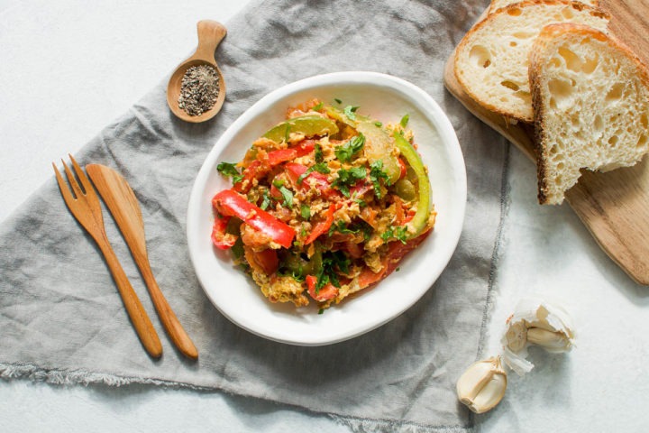 white bowl of eggs piperade on dark cloth with slices of bread to serve