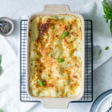 overhead photo of pesto lasagne in baking dish on cooling rack with fresh basil top right corner and baby spinach bottom left corner