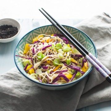 angled view of sesame noodles on blue background. Topped with spring onions and black sesame seeds ready to eat