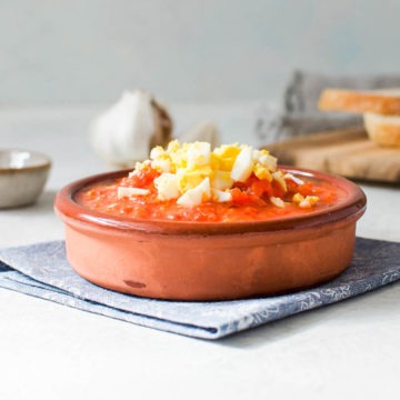side view of terracotta dish filled with roasted pepper salad topped with hardboiled egg. Slices of bread are in the background.