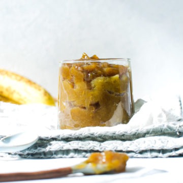 side shot of banana jam in glass on blue napkin with bananas in background