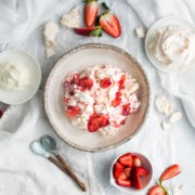 overhead shot of a bowl of eton mess with bowls of strawberries, meringue and cream round it, the background scattered with fresh sliced strawberries and broken meringue