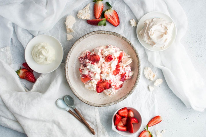 overhead shot of a bowl of eton mess with bowls of strawberries, meringue and cream round it, the background scattered with fresh sliced strawberries and broken meringue