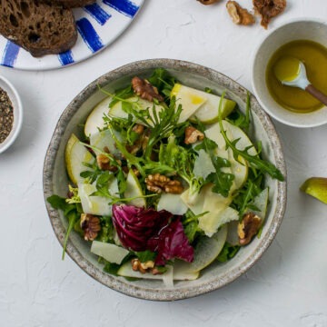one shallow bowl of walnut and pear salad with leaves and parmesan with extra lemon dressing to the right and quartered pear to the bottom of the image