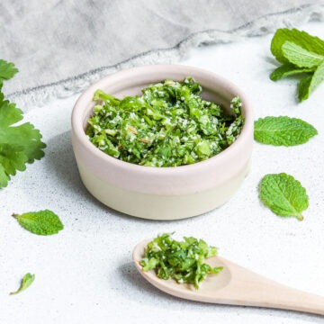 coriander chutney with coconut and mint in pink bowl and on wooden spoon with fresh mint and coriander sprinkled around