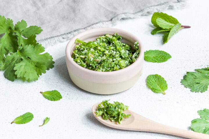 coriander chutney with coconut and mint in pink bowl and on wooden spoon with fresh mint and coriander sprinkled around