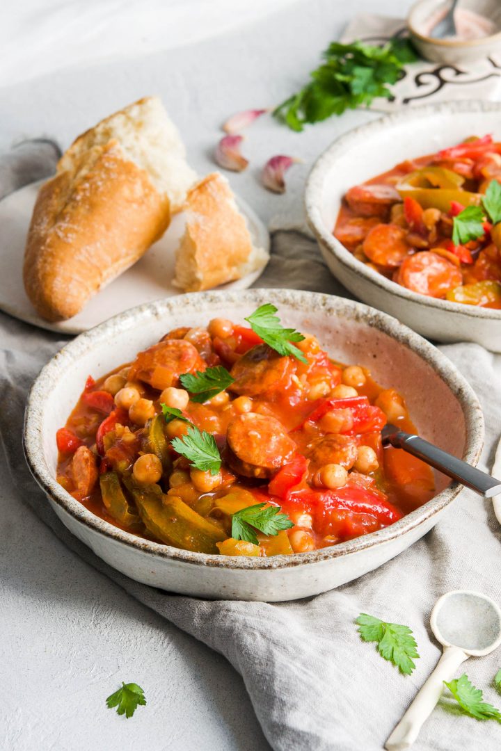 bowl of Spanish chickpea and chorizo stew sprinkled with parsley, with another bowl behind and some crusty bread to serve