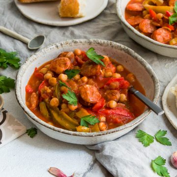 bowl of Spanish stew with chickpeas and chorizo, sprinkled with parsley and served with fresh bread which is on a plate to the right