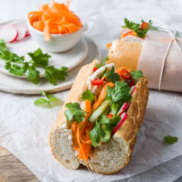 banh mi baguette with the end facing the camera, to see the variety of colourful fillings. Pickled carrot, cucumber, cold roast turkey, radish and coriander. Another banh mi sandwich is in the background, with a small bowl of extra pickled carrots