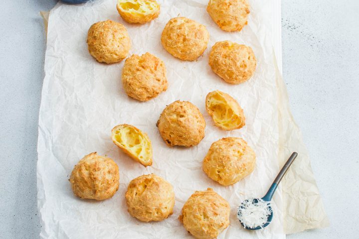French cheese puffs on white and brown baking paper, a spoon with grated cheese on the bottom right hand corner. Some of the gougeres are cut, but most are whole