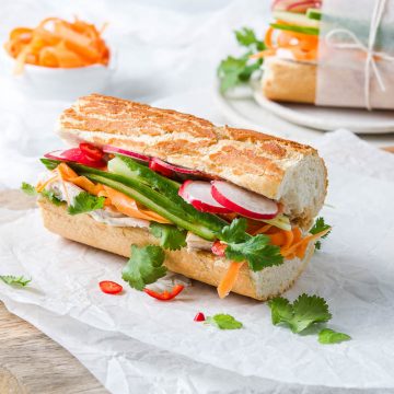 long side view of baguette filled with leftover turkey, radishes, carrot strips, fresh coriander and red chilli, with a white bowl of pickled carrot in the background together with another banh mi baguette