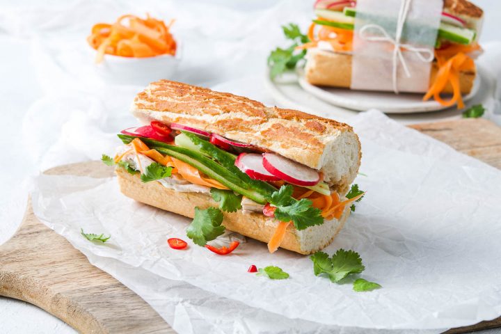 long side view of baguette filled with leftover turkey, radishes, carrot strips, fresh coriander and red chilli, with a white bowl of pickled carrot in the background together with another banh mi baguette