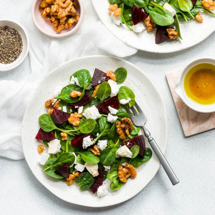 a white plate of beetroot and feta salad with spinach, extra smokey walnuts and orange dressing on the side