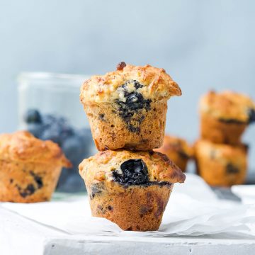 side view of two vegan blueberry muffins stacked on each other, with a glass jar of fresh blueberries in the background
