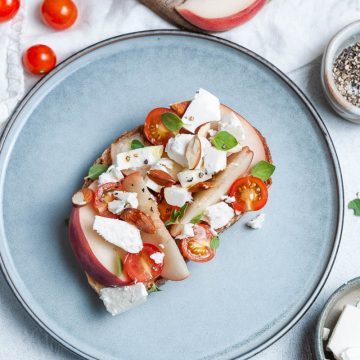 peach toast with feta, almonds, tomatoes and fresh oregano on blue plate with extra feta the bottom right of the picture, and tomatoes and white peach just seen the top of the picture