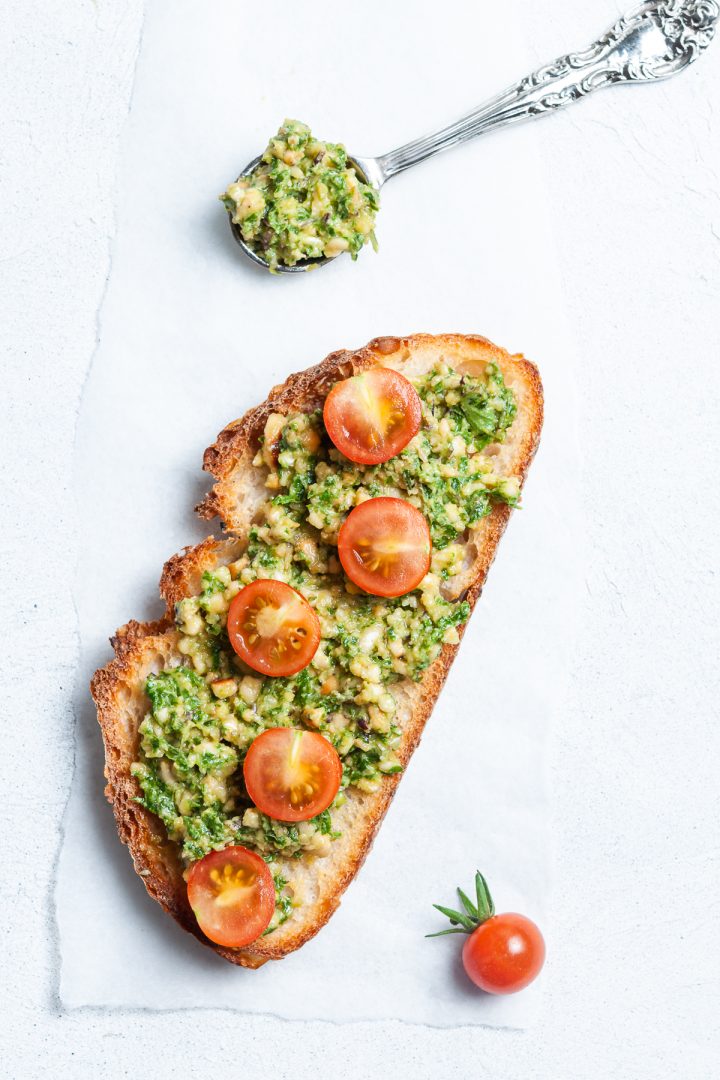 Serving suggestion: Pesto spread on a slice of toast with tomatoes on top and a spoon of extra pesto on the side.