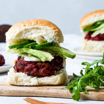 beetroot beef burger in a white bun with avocado and cucumber, on a wooden board next to a handful of rocket (arugula)