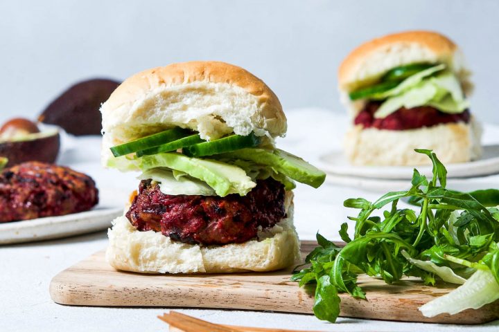 beetroot beef burger in a white bun with avocado and cucumber, on a wooden board next to a handful of rocket (arugula)