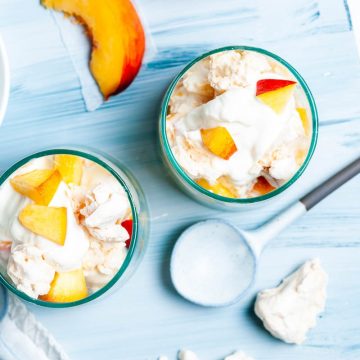 overhead shot of two glasses of peach eton mess on light blue background, with extra yellow peach slices and meringue on the board