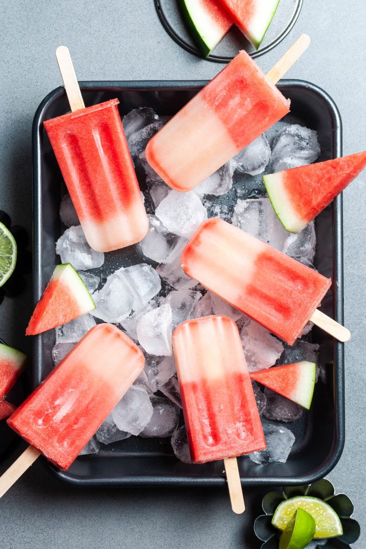 dark tray filled with ice cubers with watermelon ice lollies haphazardly arranged on top, with small wedges of fresh watermelon 