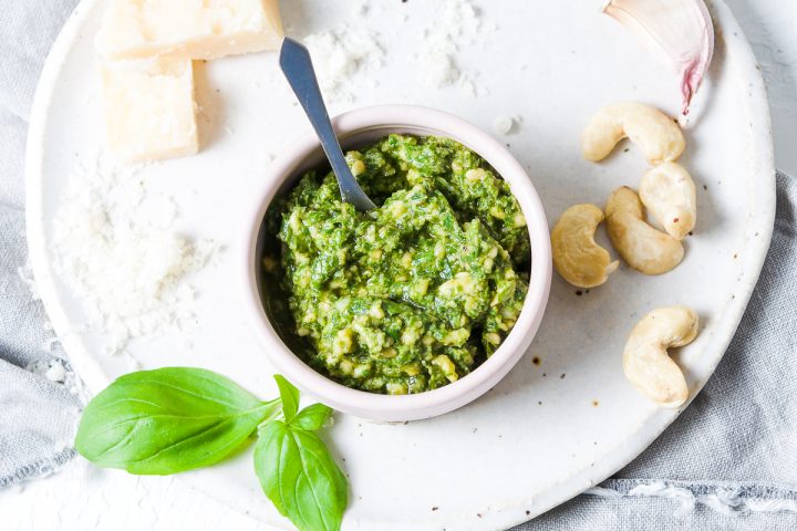 small bowl of cashew pesto on a larger plate scattered with the ingredients used in the pesto: basil, parmesan, cashew nuts and garlic
