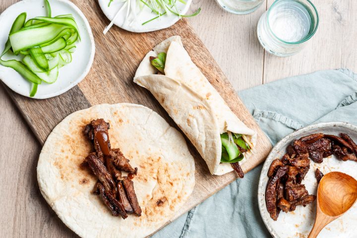 Lamb wraps being made: flatbread on a wooden board topping with hoisin lamb, cucumber and onion on a plate to be sprinkled on top. Another wrap is made to the right