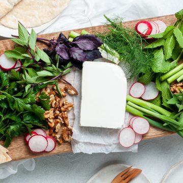 overhead image of the feta platter with a rectangle of feta cheese surrounded by fresh herbs, radishes, walnuts, and spring onions on wooden board