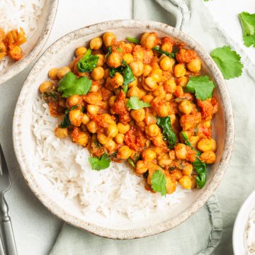 overhead photo of chickpea tomato curry in a bowl with rice, another bowl of the curry with rice just visible to the top left. Extra fresh coriander and rice are in bowls to the right