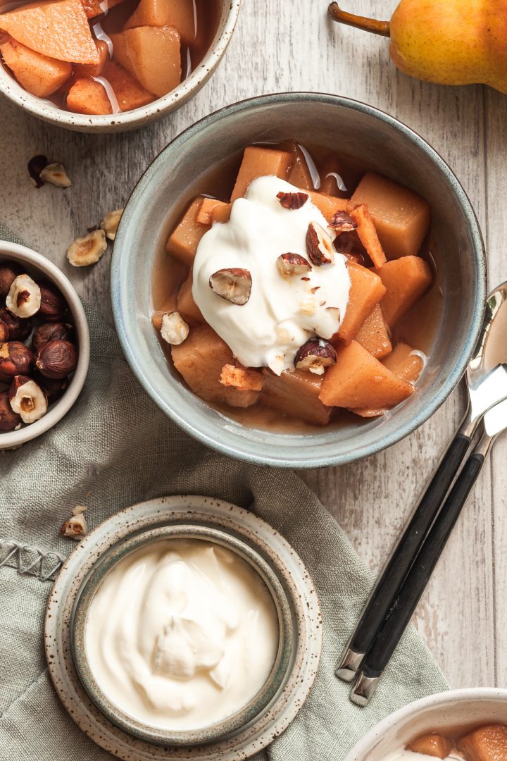 overhead shot of one bowl of cooked pears topped with plain Greek yogurt and chopped hazelnuts, a small bowl with extra yogurt is to the side