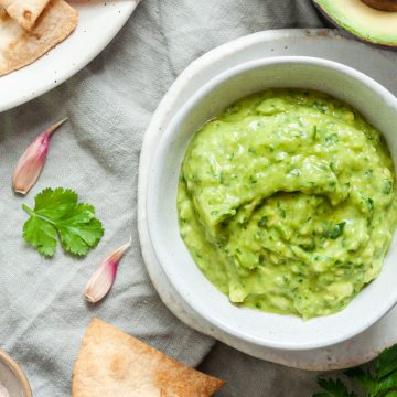 overhead view of a white bowl filled with avocado dip, with fresh coriander and garlic cloves scattered around