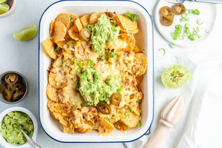 overhead photo of a full dish of tuna nachos topped with mashed avocado and chopped spring onions, lime and jalapenos at the side for those who want them on their nachos