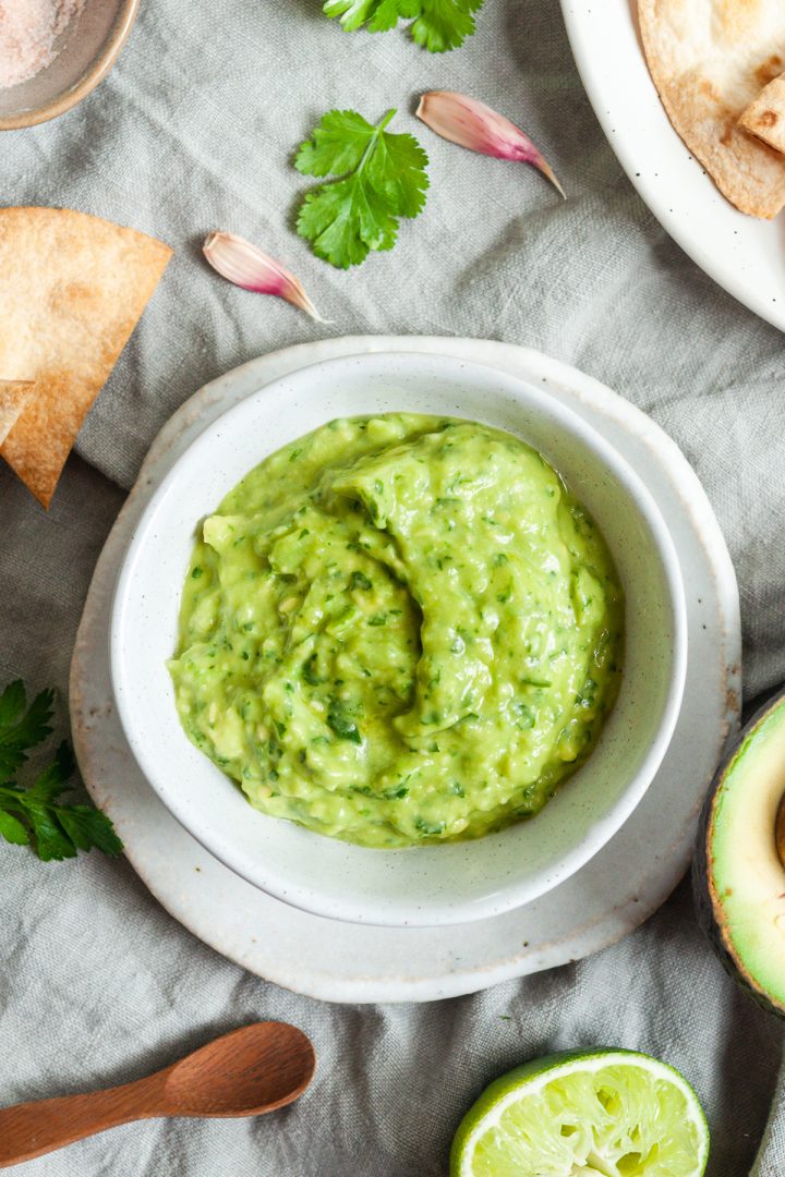 overhead photo of a bowl of avocado dip, with the main ingredients avocado, lime, garlic and coriander, dotted around the picture