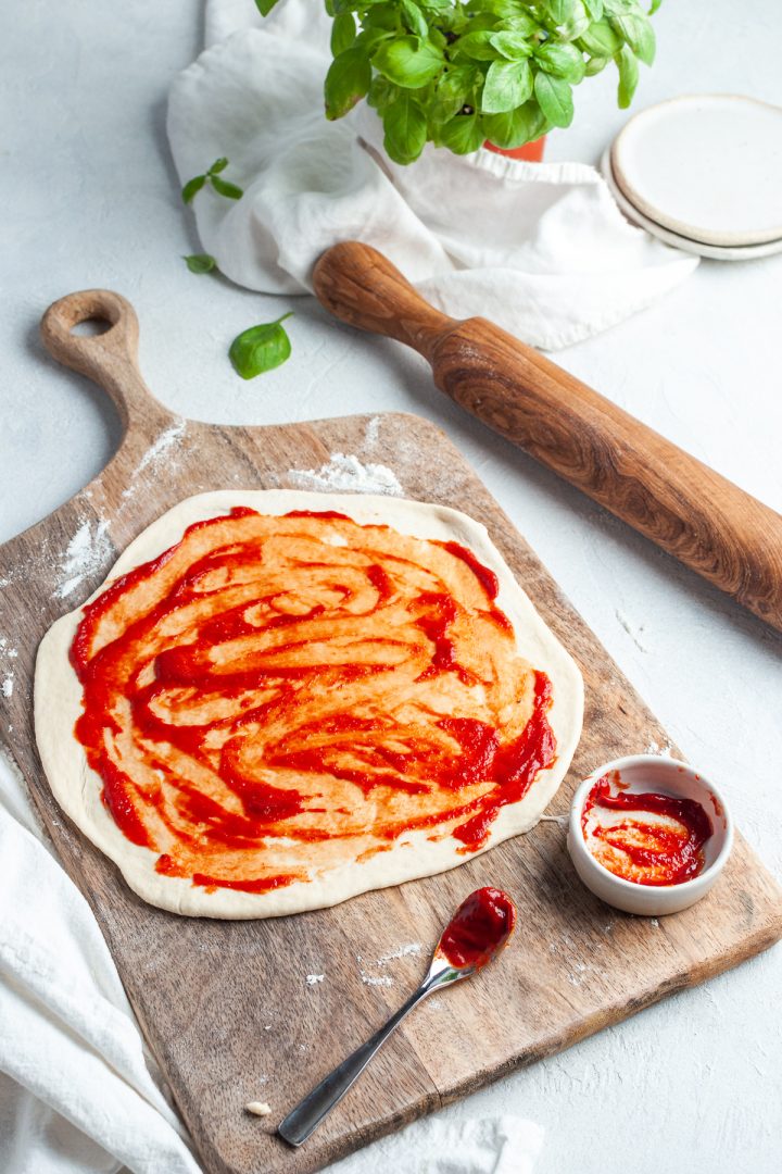 pizza dough rolled out on wooden board being spread with tomato sauce. Fresh basil ready to sprinkle on it can just be seen behind
