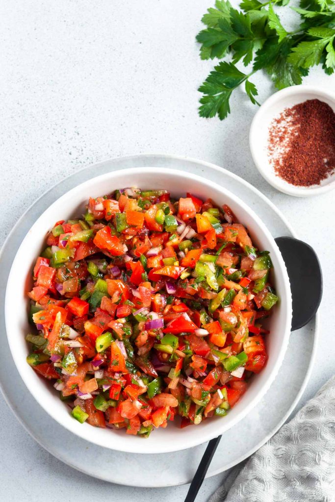 Ezme Turkish salsa in a white bowl with spoon ready to serve. A small bowl of sumac for sprinkling is to the top of the image.