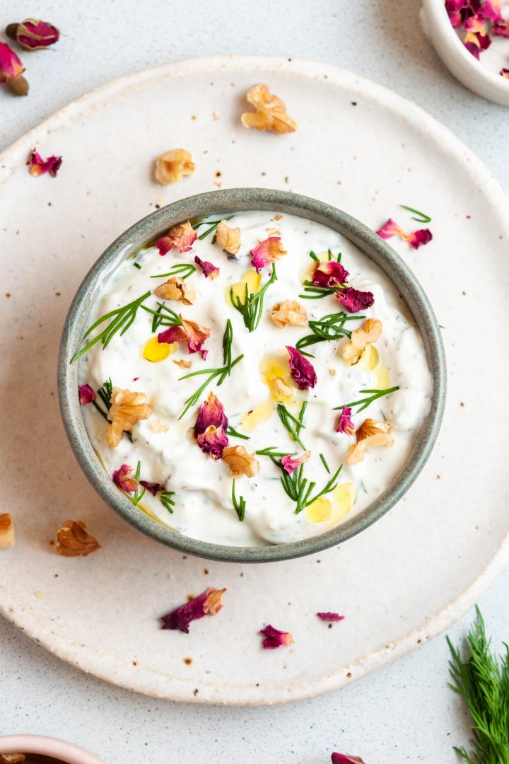 Persian yogurt dip in a bowl ready to eat, scattered with rose petals, walnuts and dill, rose petals and chunks of walnuts scattered over the background too