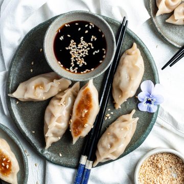 mushroom gyoza on dark blue plate next to a small bowl of dipping sauce, sesame seeds sprinkled over the top and a pair of chopsticks on the side