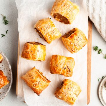 golden baked sausage rolls laid out on paper on a wooden chopping board ready to eat, fresh thyme sprinkled around
