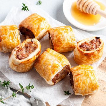 7 mini sausage rolls on white paper on wooden board ready to serve, with honey in a bowl just visible behind