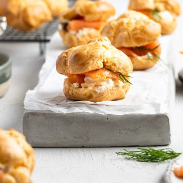 side view of a savoury profiterole filled with smoked salmon, cream cheese and fresh dill. Other filled profiteroles jut visible in the background, some on a cooling rack.