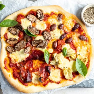overhead photo of cooked four seasons pizza on white paper on metal tray, fresh basil and cracked black pepper to the side ready to serve