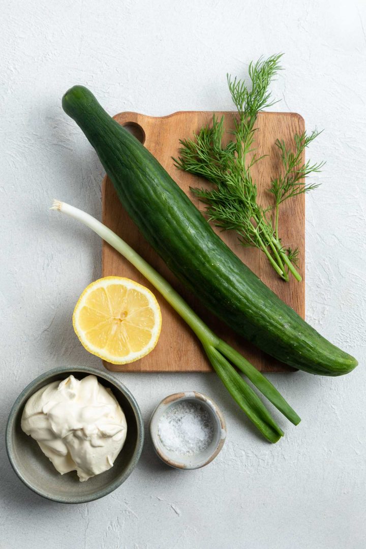 ingredients for Mizeria laid out on a wooden board: English cucumber, fresh dill, sour cream, spring onion, salt and lemon