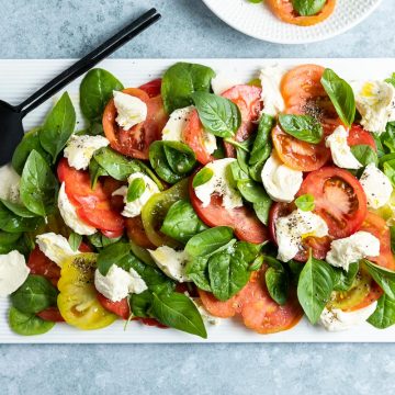 overhead photo of spinach caprese on a white rectangular plate, black serving spoons on the top left corner ready to serve