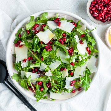 overhead photo of rocket pomegranate salad in a bowl on a white cloth, a small bowl of pomegranate seeds and another bowl of vinaigrette to the side