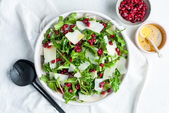overhead photo of rocket pomegranate salad in a bowl on a white cloth, a small bowl of pomegranate seeds and another bowl of vinaigrette to the side